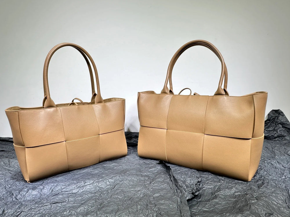 Two tan woven leather tote bags with sturdy handles displayed on crumpled black fabric.