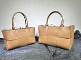 Two tan woven leather tote bags with sturdy handles displayed on crumpled black fabric.