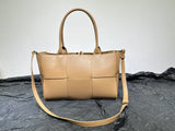 Tan woven leather tote bag with top handles and adjustable shoulder strap on textured black surface