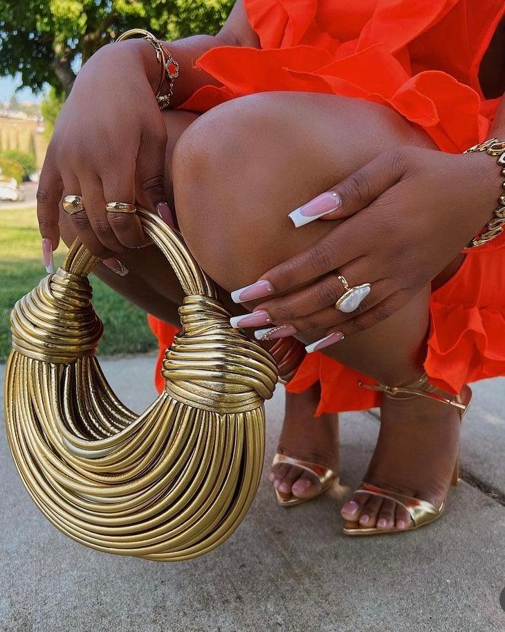 Woman holding a gold leather designer handbag, wearing an orange dress, gold heels, and jewelry