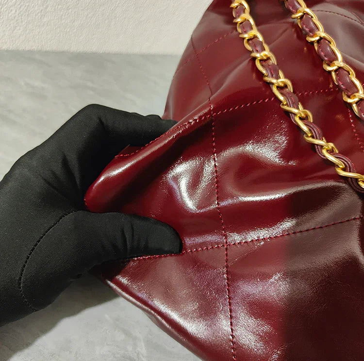 Close-up of quilted burgundy leather shoulder bag with gold chain strap, held by gloved hand.