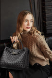 Woman holding black woven leather shoulder bag with metal buckle, wearing brown knit sweater.