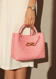 Pink woven leather shoulder bag with gold metal buckle, held by woman in white dress