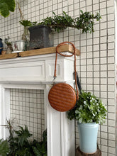 Round woven leather crossbody bag with braided strap hanging on a white mantel, surrounded by potted plants indoors.
