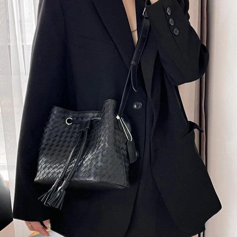 Handwoven black lambskin leather bucket bag with drawstring, crossbody style, worn by a woman in a black blazer