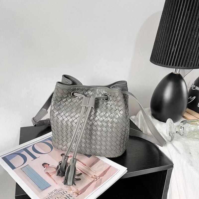 Gray handwoven lambskin leather bucket bag with drawstring and tassels on table