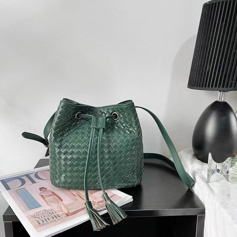 Green handwoven lambskin leather bucket bag with drawstring and tassels on a table.