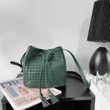Green handwoven lambskin leather bucket bag with drawstring and tassels on a table.