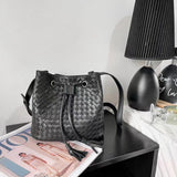 Handwoven black lambskin leather bucket bag with drawstring closure on a table next to a magazine and lamp