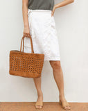 Woman holding a hand-woven brown rattan tote bag, styled with white wrap skirt and sandals