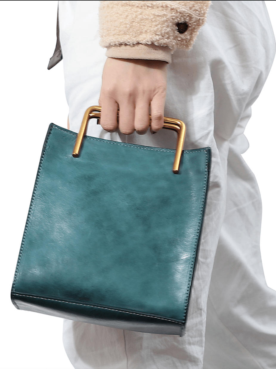 Teal cowhide leather shoulder bag with gold metal handle, held by woman in white pants