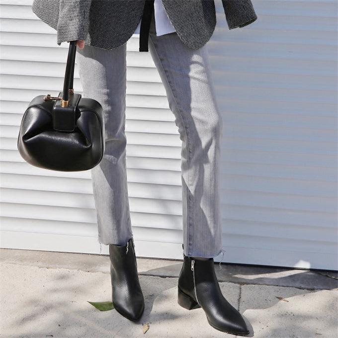 Woman holding black genuine leather round handbag with gold lock, wearing grey pants and black boots
