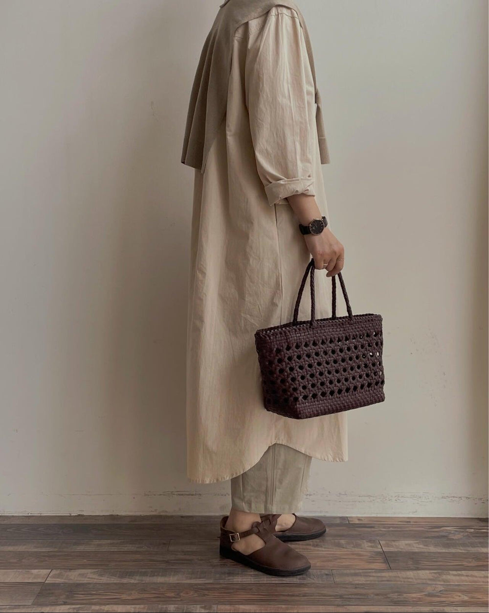 Woman holding a brown handwoven leather tote bag, wearing neutral dress and brown shoes