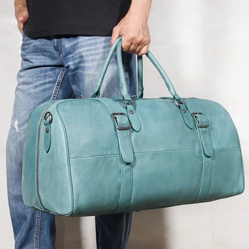 Hand holding a teal full-grain leather duffle bag with handles and buckled straps
