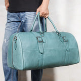 Hand holding a teal full-grain leather duffle bag with handles and buckled straps