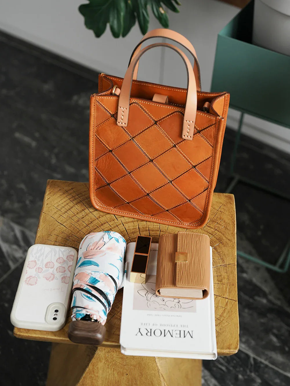 Handcrafted brown cowhide leather handbag with woven pattern on wooden table, surrounded by wallet, floral umbrella, phone, lipstick, and notebook.