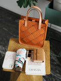 Handcrafted brown cowhide leather handbag with woven pattern on wooden table, surrounded by wallet, floral umbrella, phone, lipstick, and notebook.