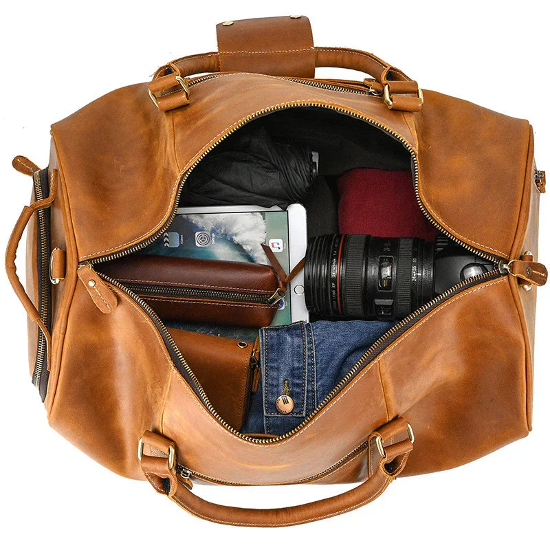 Open brown full-grain leather duffle bag with clothing, camera, tablet, and travel essentials inside