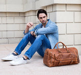 Handmade full grain leather duffle bag in dark brown with shoe compartment, beside a seated man in casual denim attire outdoors.