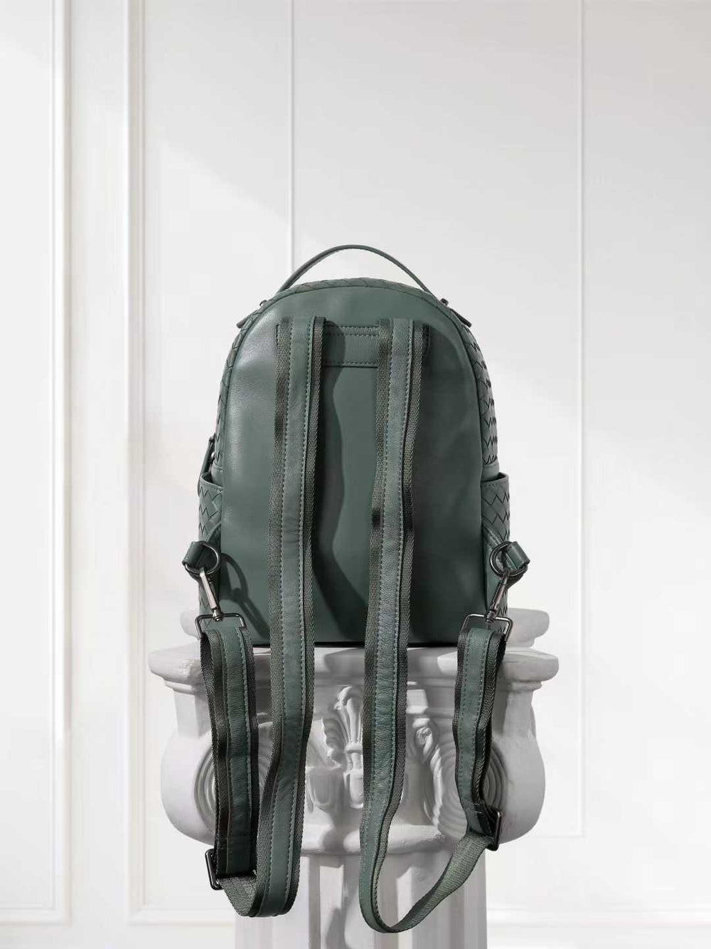 Back view of a small green lambskin leather backpack with woven side details on a pedestal.
