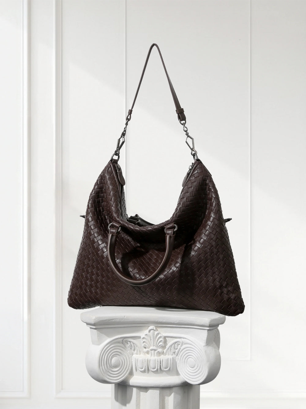Large black woven lambskin leather tote bag with dual handles and shoulder strap, displayed on a white pedestal.
