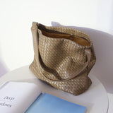 Beige woven leather tote bag with shoulder strap on a white table beside an open book