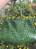 Green woven leather hobo bag with double handles, displayed outdoors among yellow flowers