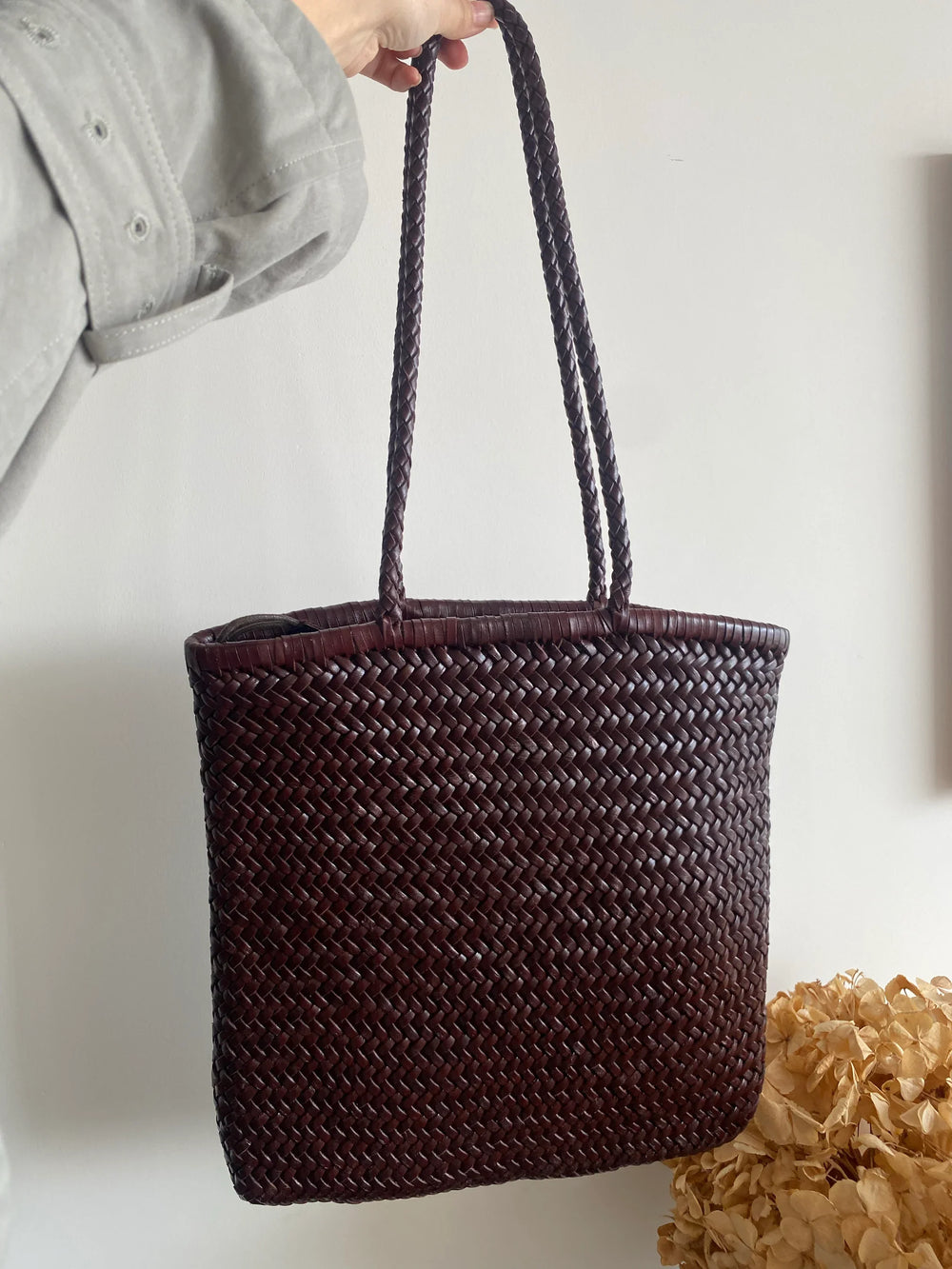 Hand holding a dark brown woven leather tote bag with braided handles, against a neutral background.