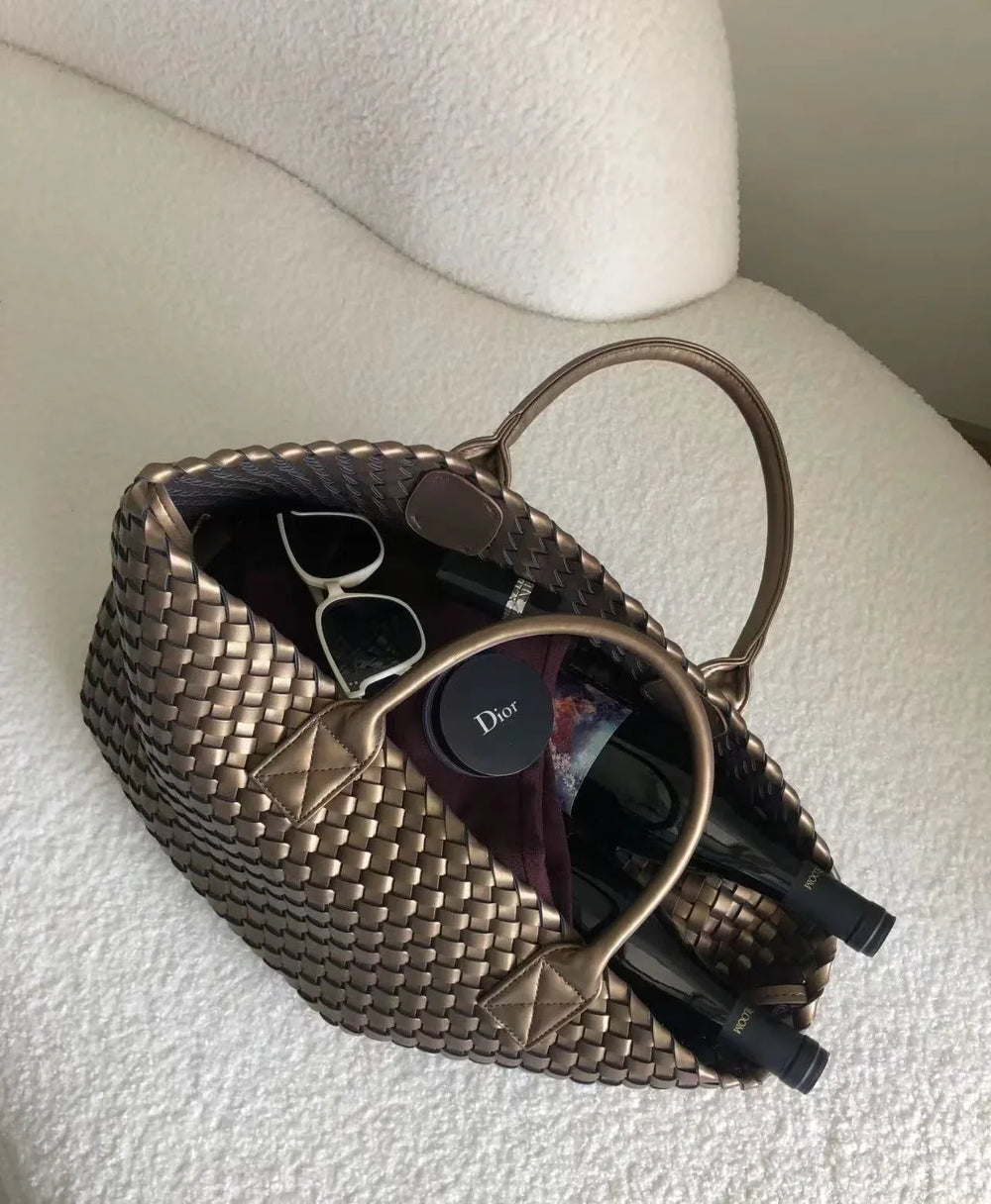 Large handwoven vegan leather tote bag in onyx filled with sunglasses, wine bottles, and accessories, displayed on a textured white chair.