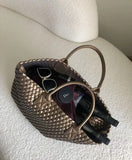 Large handwoven vegan leather tote bag in onyx filled with sunglasses, wine bottles, and accessories, displayed on a textured white chair.