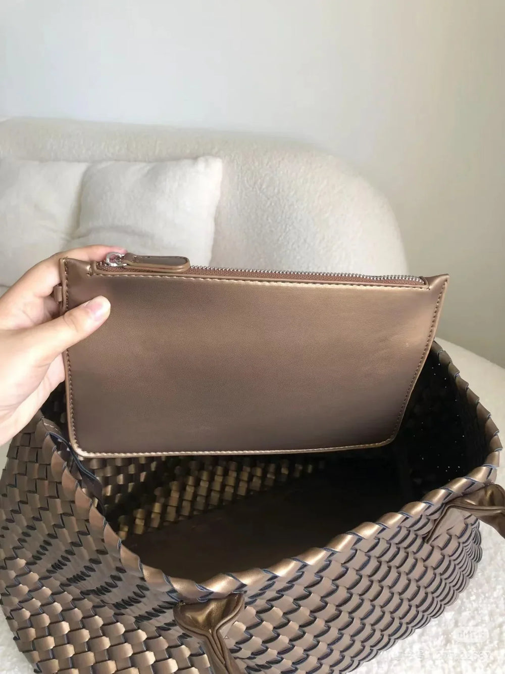 Hand holding brown pouch above large woven vegan leather tote bag on neutral sofa