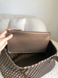 Hand holding brown pouch above large woven vegan leather tote bag on neutral sofa