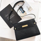 Black leather crossbody bag with crocodile pattern and gold hardware on a white table.