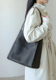 Minimalist dark grain cowhide leather tote bag with shoulder strap, worn by woman in light trench coat