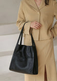 Minimalist black grain cowhide leather tote bag with double shoulder straps, held by woman in beige suit
