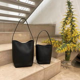 Two minimalist black leather bucket bags with shoulder straps on stone stairs next to yellow flowers.