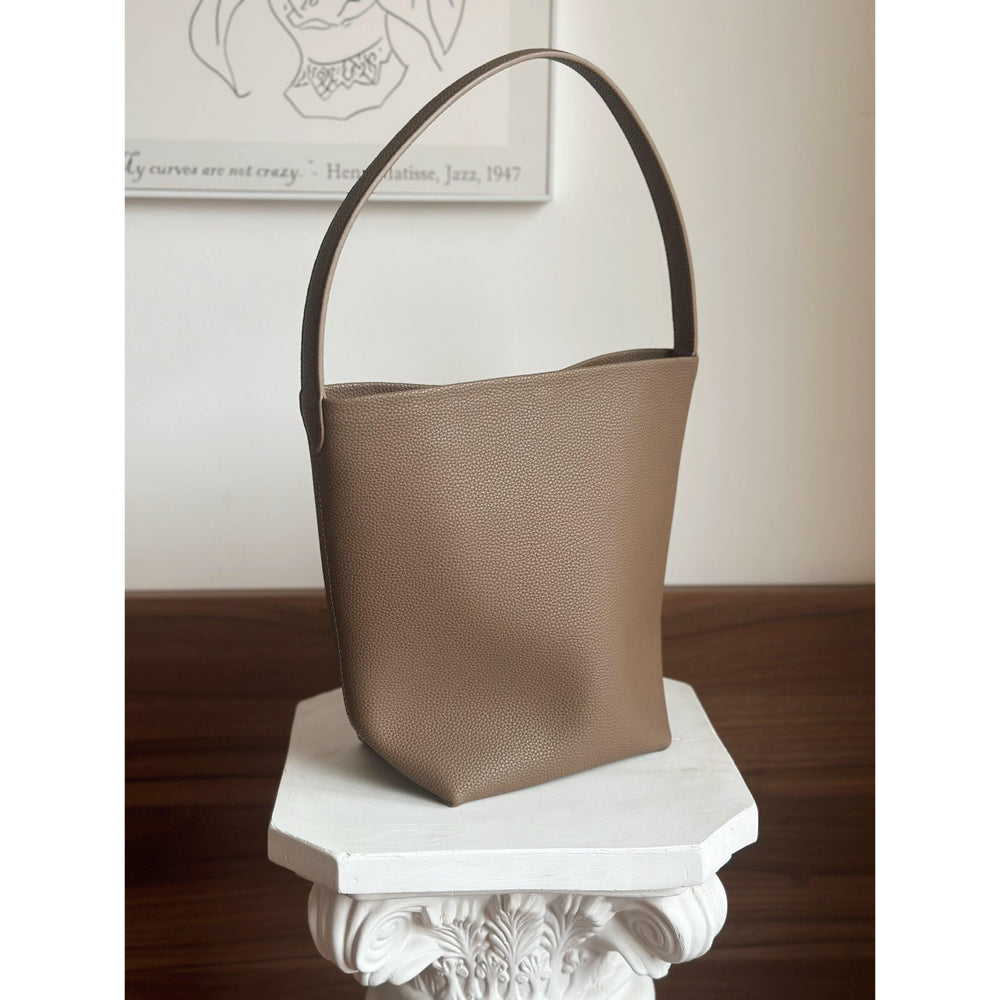Minimalist tan leather bucket bag with single shoulder strap on white pedestal