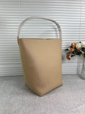 Minimalist tan leather bucket bag with wide handle on grey surface near flower vase