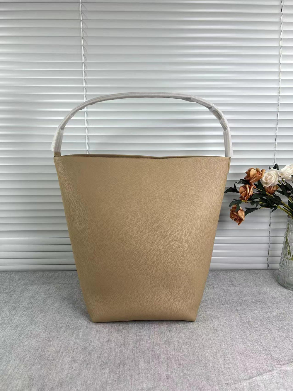 Minimalist tan leather bucket bag with sturdy handle on a grey surface, flowers in background