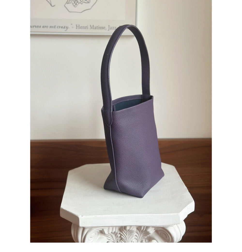 Minimalist purple leather bucket bag with shoulder strap on decorative white pedestal