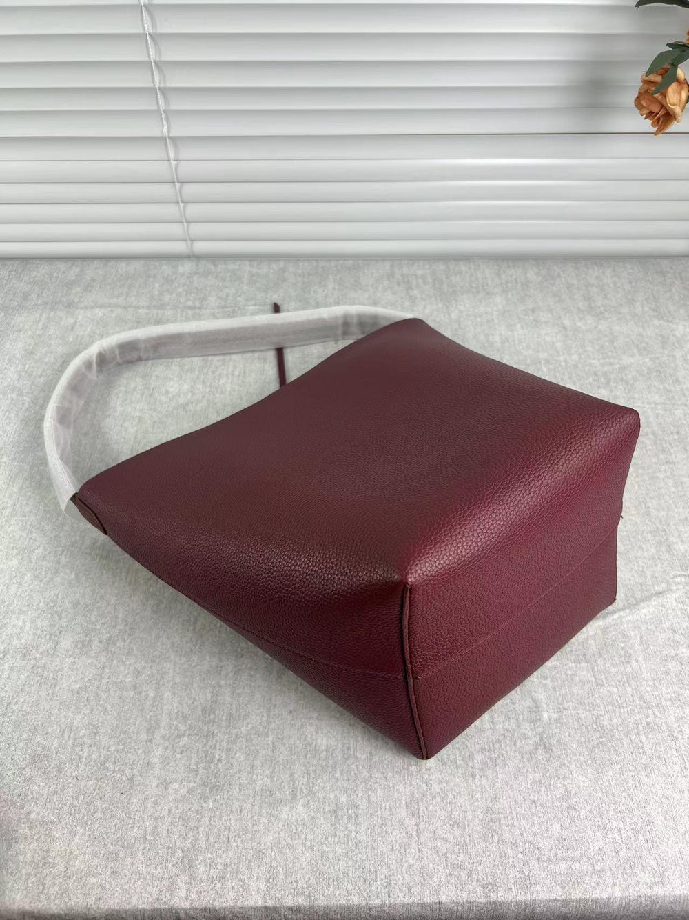 Minimalist burgundy leather bucket bag with shoulder strap on a light fabric surface