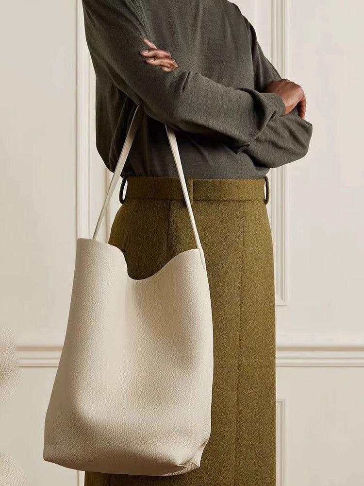 Minimalist tan leather bucket bag with shoulder strap, worn by person in neutral outfit.