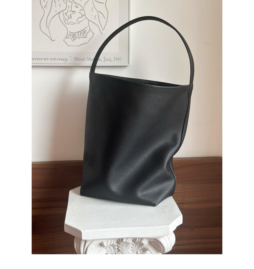 Minimalist black leather bucket bag with single strap on white pedestal, modern background