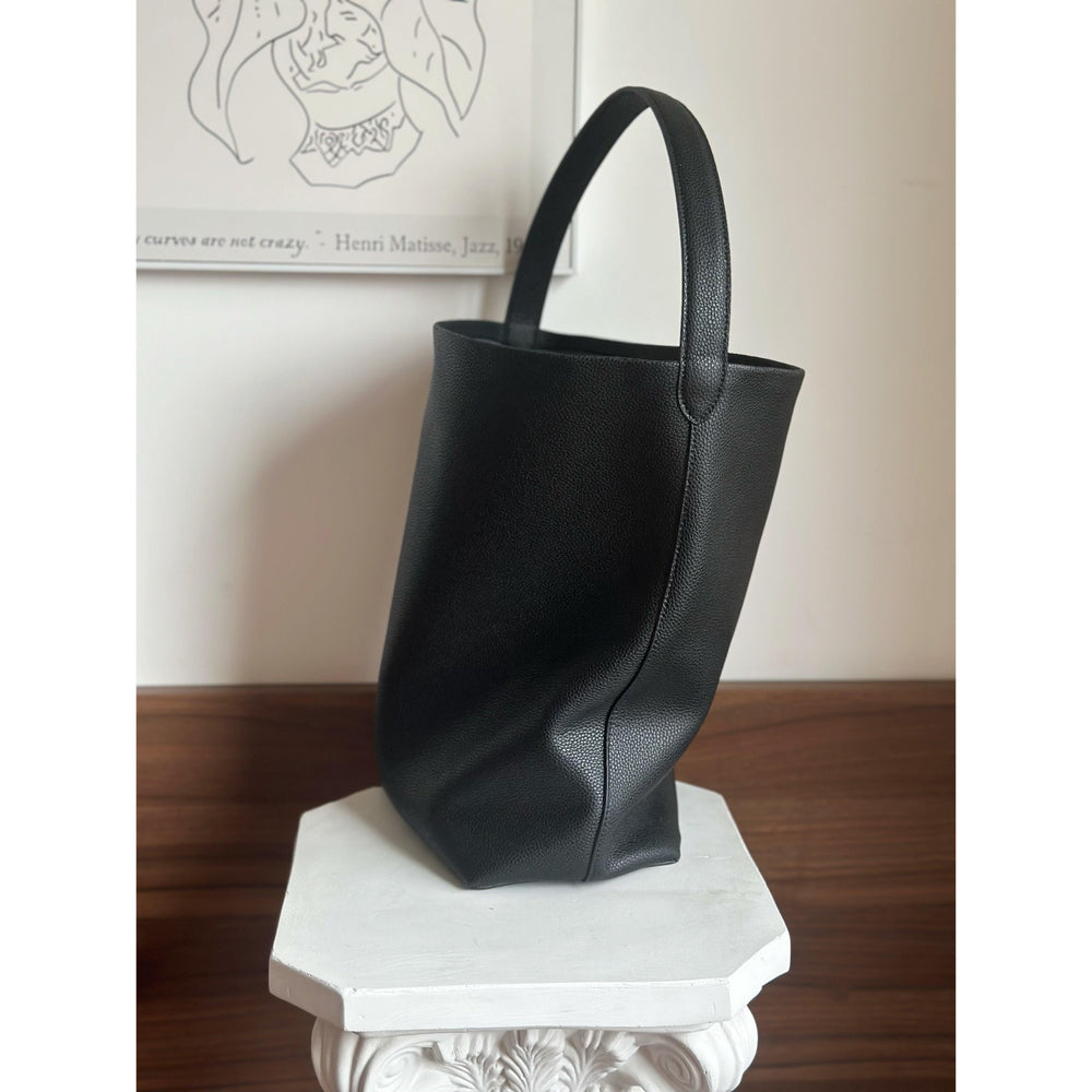 Minimalist black leather bucket bag with single shoulder strap on a white pedestal indoors