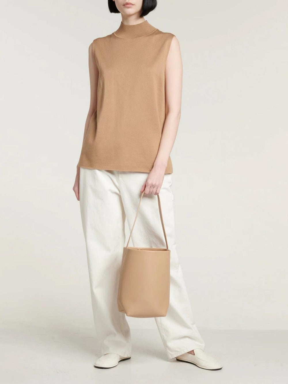 Woman holding minimalist tan leather bucket bag, styled with white pants and beige top
