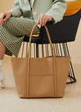 Minimalist beige genuine leather tote bag with top handles, shown on a seated woman indoors.