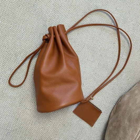Minimalist brown leather drawstring bucket bag with shoulder strap on textured surface