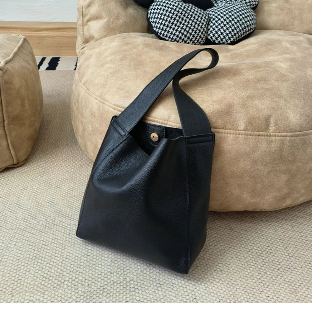 Minimalist black pebbled cowhide leather bucket bag with wide shoulder strap on beige pouf