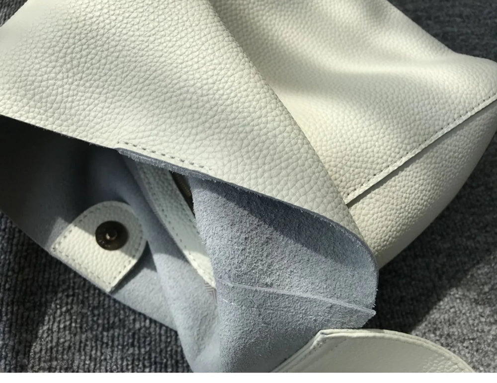 Minimalist pebbled cowhide leather bucket bag with wide shoulder strap on gray surface