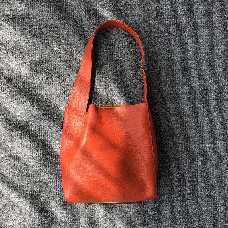 Minimalist orange pebbled cowhide leather bucket bag with wide shoulder strap on gray fabric
